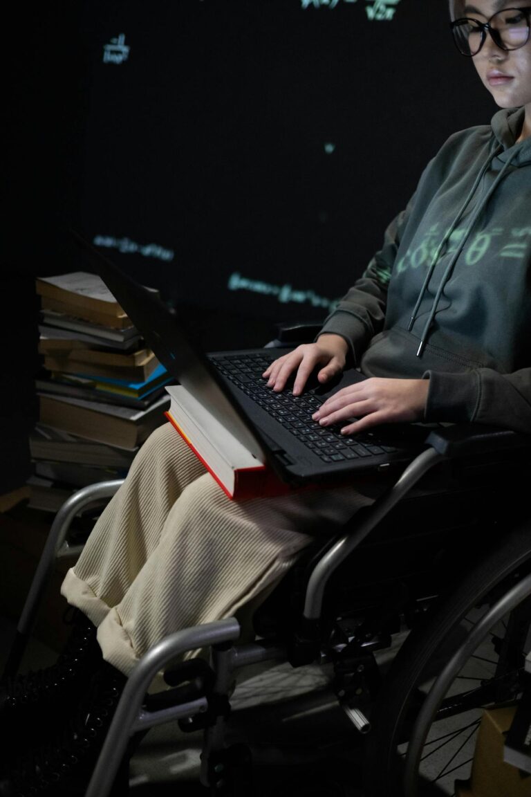 A teenager in a wheelchair typing on a laptop, surrounded by books, suggesting studying.