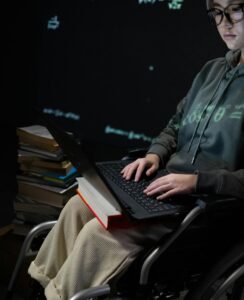 A teenager in a wheelchair typing on a laptop, surrounded by books, suggesting studying.