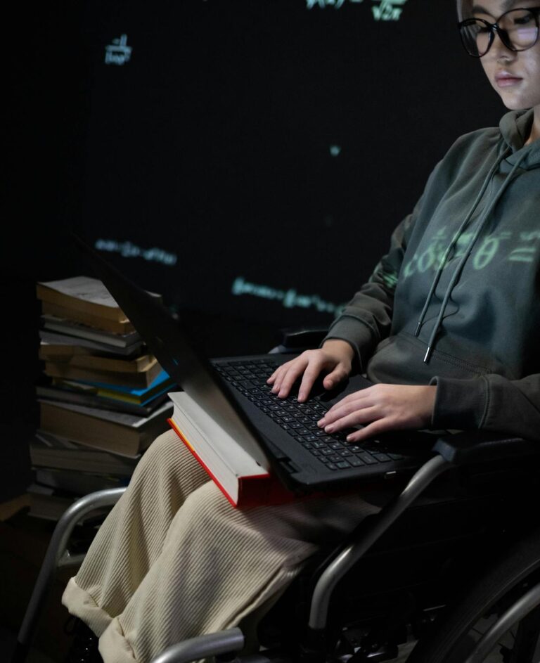 A teenager in a wheelchair typing on a laptop, surrounded by books, suggesting studying.