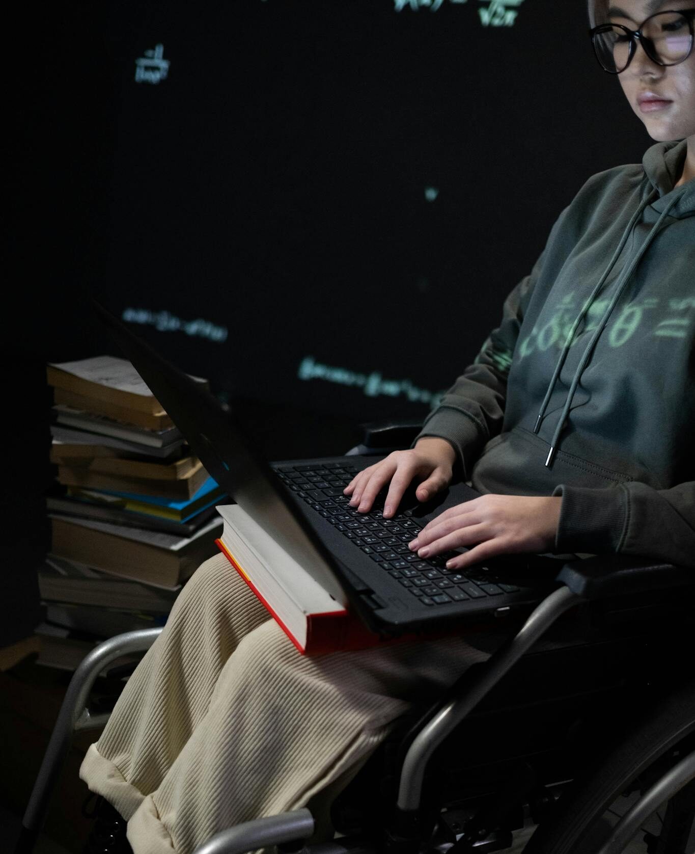 A teenager in a wheelchair typing on a laptop, surrounded by books, suggesting studying.