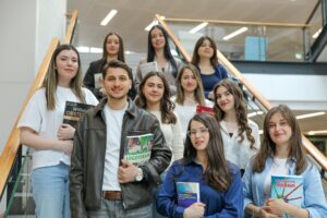 Diverse group of students smiling on stairs, holding educational books indoors.